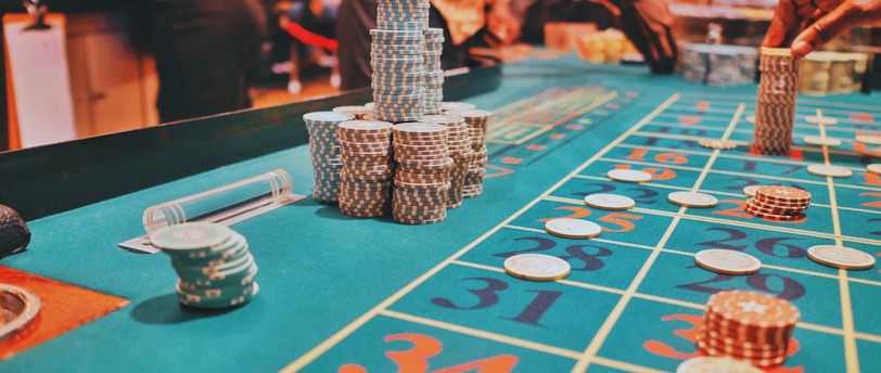A casino scene with a roulette table features prominently, covered in green felt with colorful numbers and betting squares. Several stacks of poker chips are placed on different numbers, and the hands of several people are visible as they interact with the table. In the background, there are slot machines and more people, creating a vibrant and busy atmosphere.