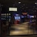 A dimly lit casino with several rows of slot machines, some displaying bright graphics and lights. There are a few people scattered throughout the area, engaging with the machines. The floor is reflective and polished, adding to the ambiance, while the ceiling is dotted with numerous small lights.
