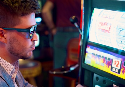 A man wearing sunglasses is intently focused on a slot machine, with colorful lights and graphics illuminating the screen. The scene is set indoors, likely in a casino or gaming area. His attire includes a patterned shirt and a blazer.