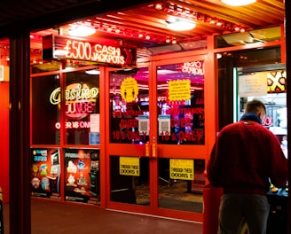A brightly lit entrance to a casino with neon signs, including one advertising a £500 jackpot. The doors have clear signage indicating entry is restricted to those over 18. A person in a red jacket stands to the right, possibly interacting with a machine or display.